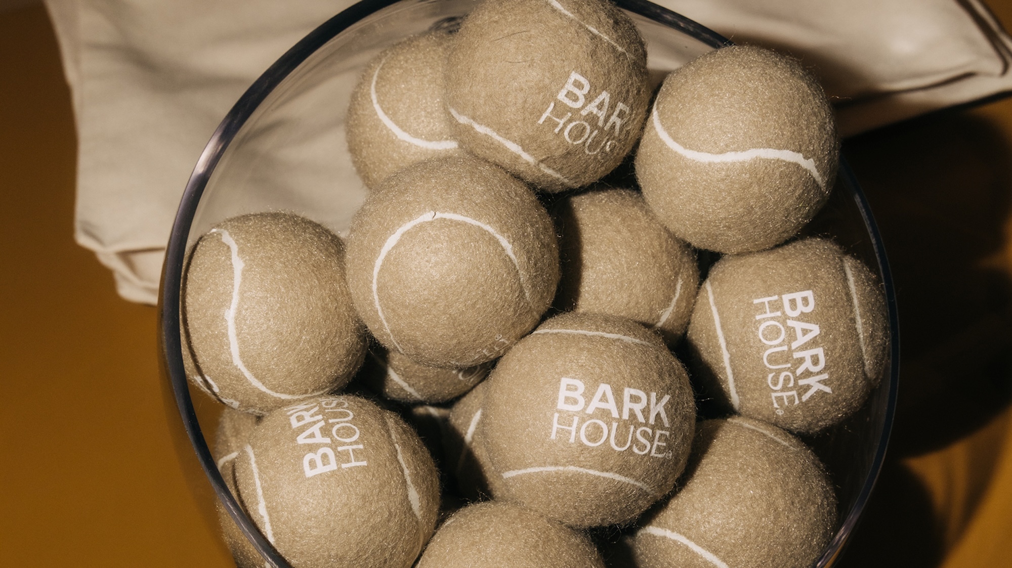 Tan tennis balls with white Barkhouse branding piled inside a clear glass container against a warm background.
