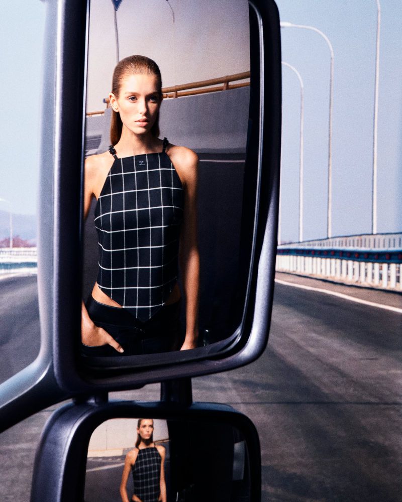 Model reflected in a vehicle side mirror wearing a black grid-patterned halter top on an elevated roadway