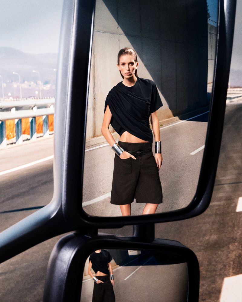 Model reflected in a truck side mirror wearing a black draped top and shorts with wide silver cuffs against an asphalt road backdrop.