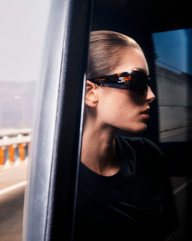 Profile of a woman wearing dark tortoiseshell sunglasses and a black top inside a vehicle looking out.