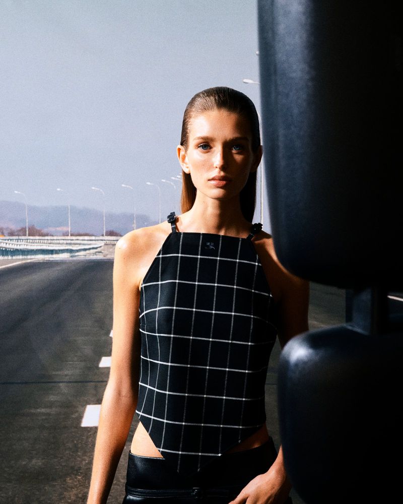 Model wearing a black checkered halter top outdoors near a highway with a blurred dark object in the foreground.