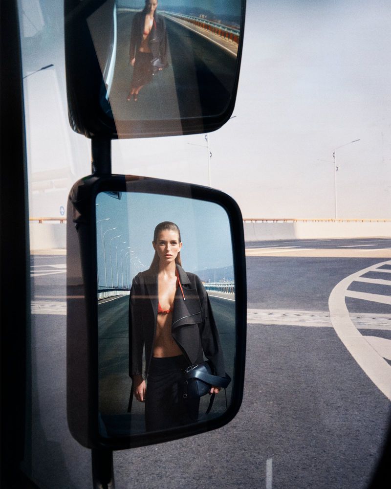 Model reflected in a truck side mirror standing on a modern overpass roadway wearing a black jacket and carrying a handbag