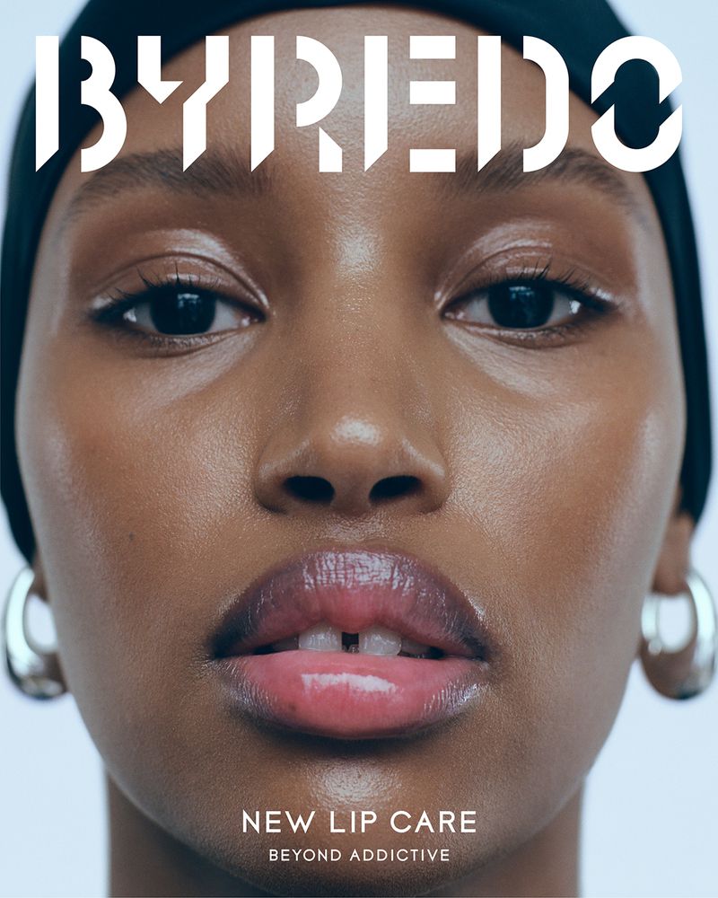 Close-up of an adult female model of African descent with dark hair, wearing a black head covering and silver hoop earrings. Makeup focuses on lips.