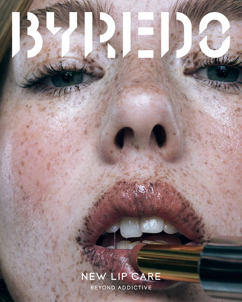 Close-up shot of a fair-skinned adult woman with freckles applying lipstick with a gold tube. Brand logo, 'BYREDO,' in view.