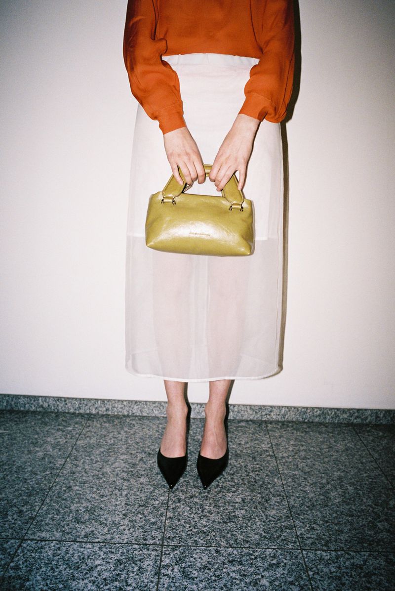 Model wearing a rust-orange top, white sheer skirt, and black pointed heels holding a green handbag