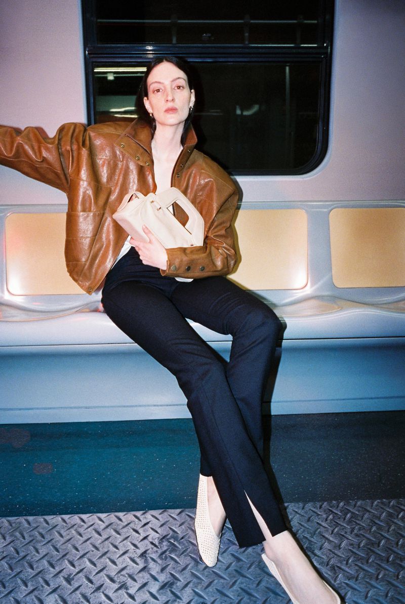 Model wearing a brown leather jacket and dark trousers holds a light handbag while seated inside a train carriage.