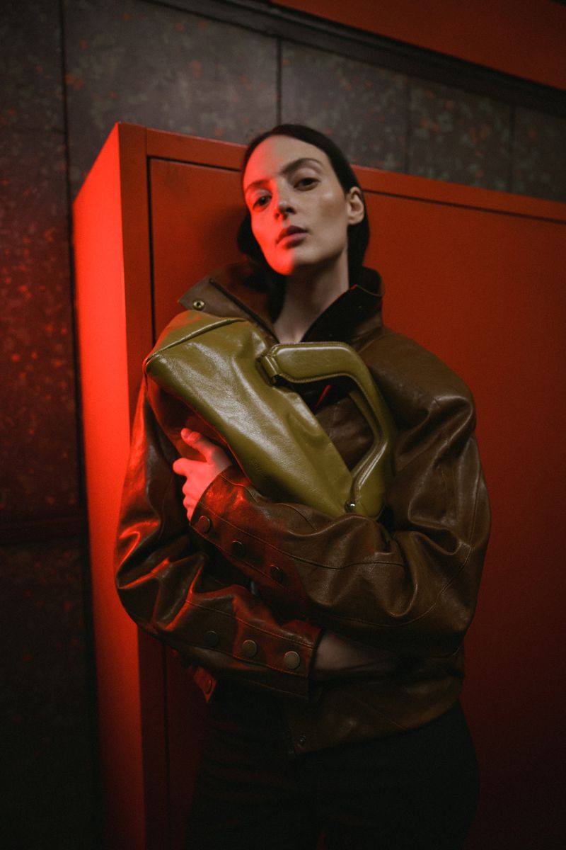Model wearing a brown leather jacket holding an olive green handbag against an orange locker backdrop