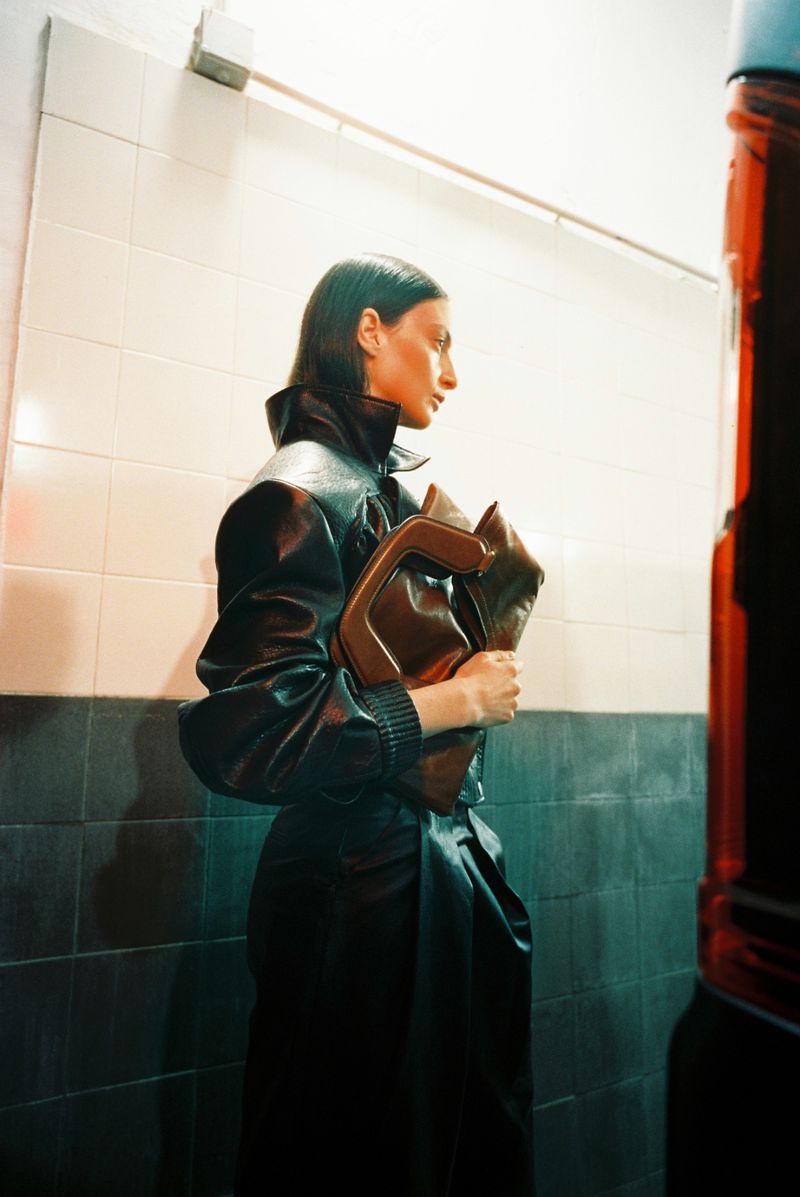 Model wearing dark leather jacket holding brown structured handbag against tiled wall for AIM SS 26 campaign