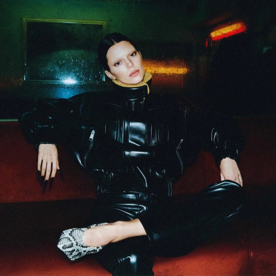 A woman in black leather attire poses indoors with snake-print shoes, against a backdrop of dark colors and muted lighting.