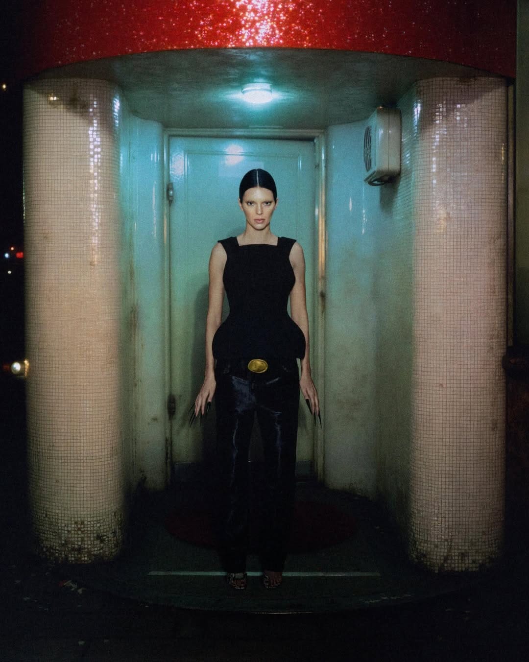 Model Kendall Jenner wearing a black top and dark trousers standing symmetrically between tiled columns for Khaite.