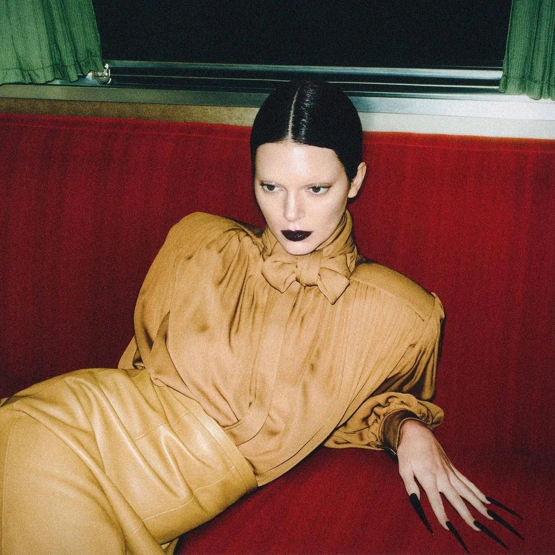 Kendall Jenner lounging on a vibrant red tufted banquette wearing a camel blouse with long pointed black nails.