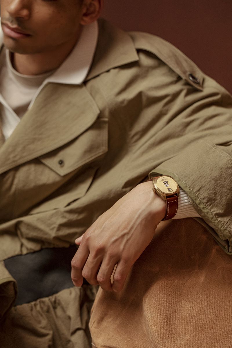 Tan-skinned man in trench coat poses indoors with wrist watch. Focus on outfit and accessory in studio setting with warm lighting.