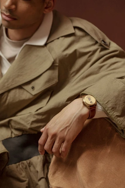 Tan-skinned man in trench coat poses indoors with wrist watch. Focus on outfit and accessory in studio setting with warm lighting.