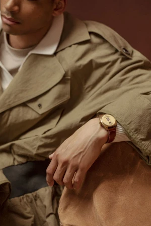Tan-skinned man in trench coat poses indoors with wrist watch. Focus on outfit and accessory in studio setting with warm lighting.