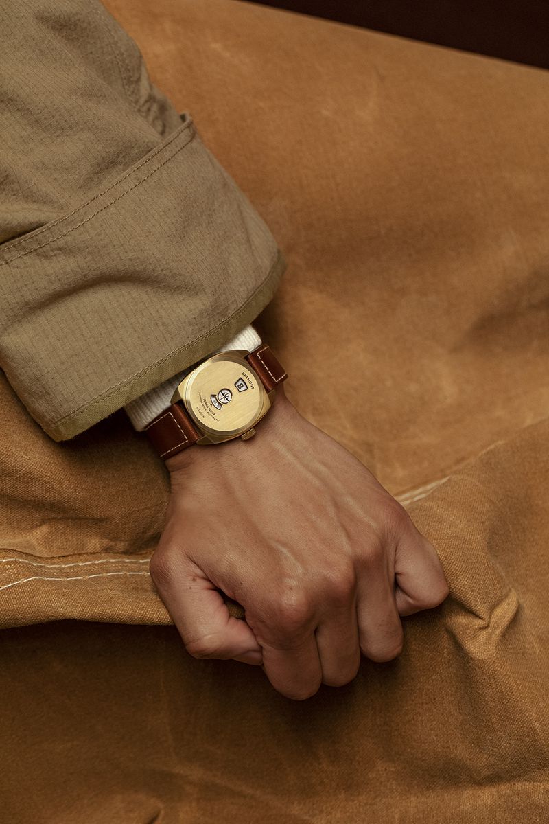 Close-up of a person's hand wearing a square gold watch with a brown leather strap, set against a tan textured background.