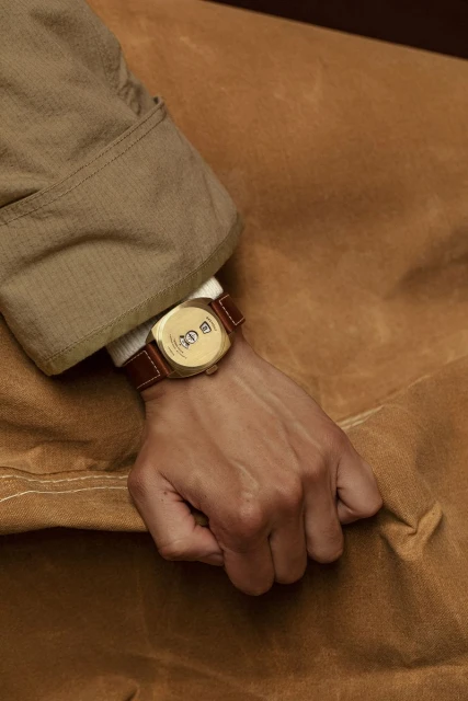 Close-up of a person's hand wearing a square gold watch with a brown leather strap, set against a tan textured background.