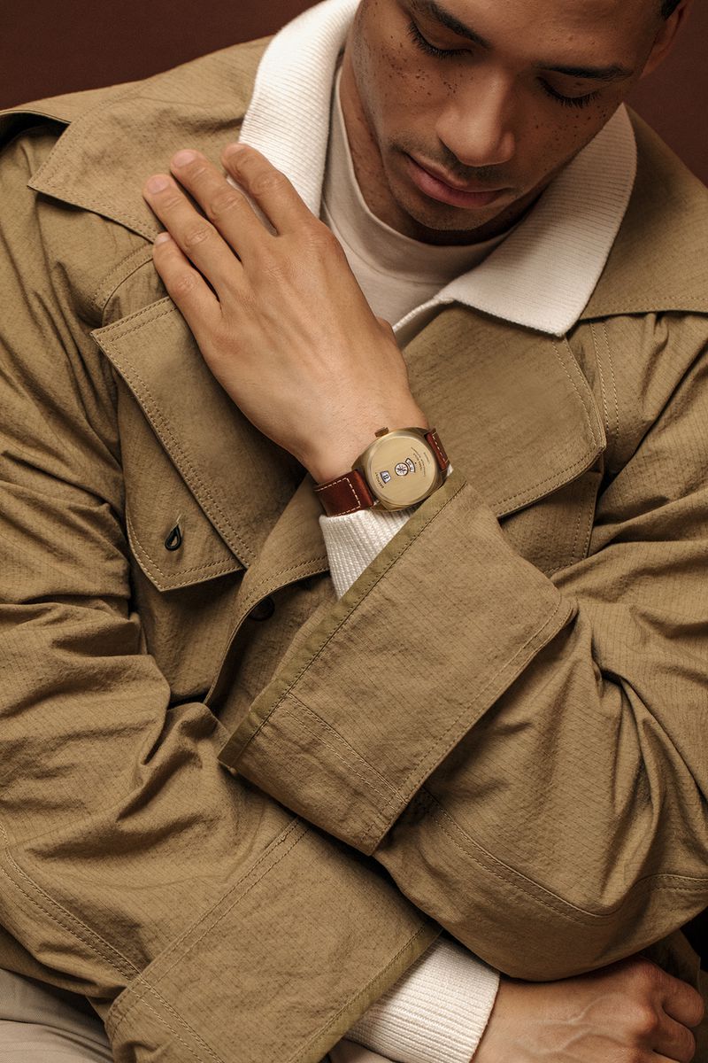 Adult male in trench coat showcases brown wristwatch with brown leather band against a dark brown backdrop in a studio shot.