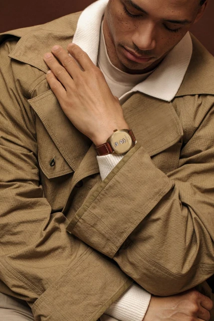 Adult male in trench coat showcases brown wristwatch with brown leather band against a dark brown backdrop in a studio shot.