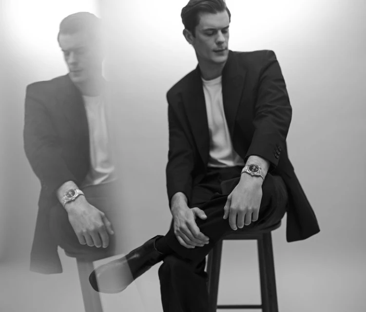 Black and white portrait of an adult male in a dark suit and light shirt, sitting on a stool and wearing a wristwatch.