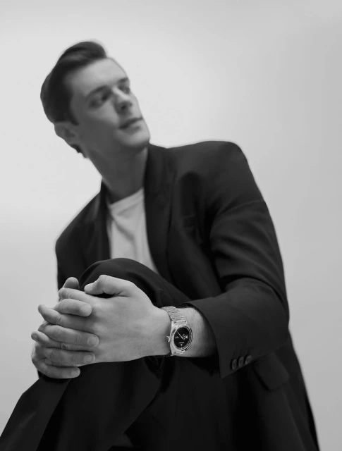 Black and white portrait of an adult male model wearing a suit, with a watch, and kneeling indoors.