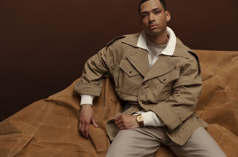 A light-skinned man poses indoors wearing a tan coat, sweater, and pants, sitting on a draped fabric in a studio setting with brown backdrop.