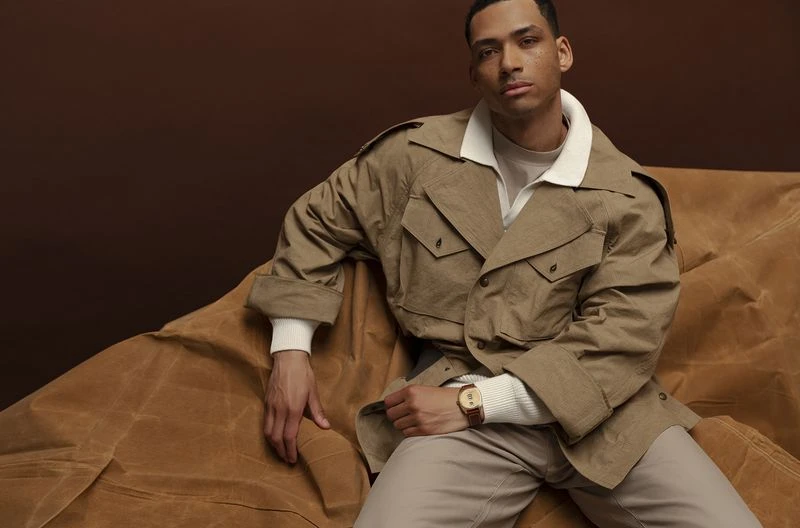A light-skinned man poses indoors wearing a tan coat, sweater, and pants, sitting on a draped fabric in a studio setting with brown backdrop.
