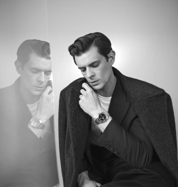 Monochrome portrait of a dark-haired man in a suit and coat, looking at his wristwatch with a reflection in a glass panel.