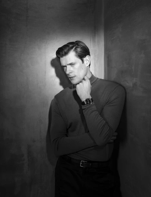 Black and white portrait of an adult male with styled dark hair, wearing a turtleneck and watch, posing against a wall.