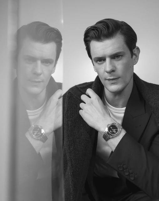 Black and white portrait of an adult male wearing a suit coat, with a wristwatch, and a reflection, in an indoor setting with diffused light.