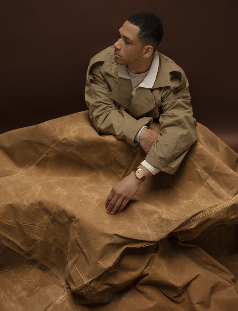 Studio shot of a man wearing a tan coat, posing against a dark brown background with tan fabric draped around him. The man is looking to the side.