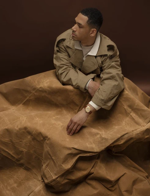 Studio shot of a man wearing a tan coat, posing against a dark brown background with tan fabric draped around him. The man is looking to the side.