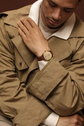 Model Lewis Jamison wearing a tan jacket and a gold jumping hour watch with a brown leather strap against a brown studio background.