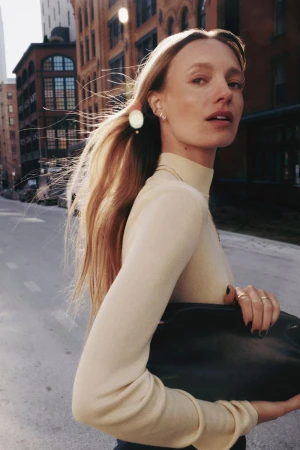 Model with long blonde hair in a cream turtleneck holding a black handbag walking on a city street.