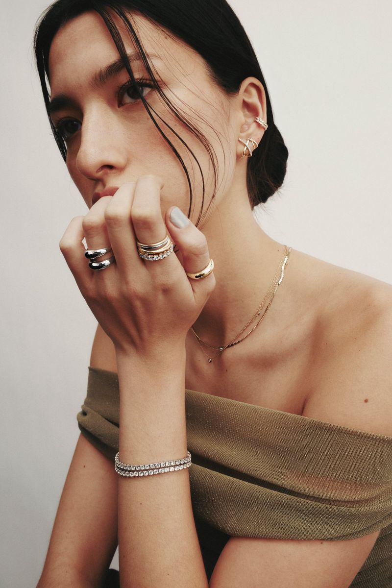 Close-up portrait of a model wearing stacked rings, layered necklaces, and bracelets against a plain light background for Mejuri.