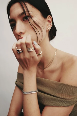 Close-up portrait of a model wearing stacked rings, layered necklaces, and bracelets against a plain light background for Mejuri.