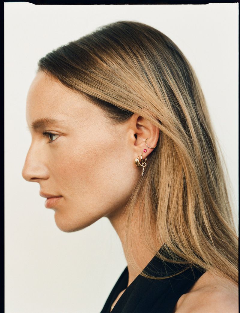 Profile portrait of a woman wearing gold hoop earrings and delicate jewelry against a plain light background.