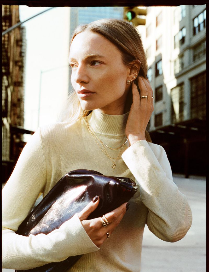 Woman wearing gold jewelry and a pale yellow turtleneck holding a dark clutch on a city street