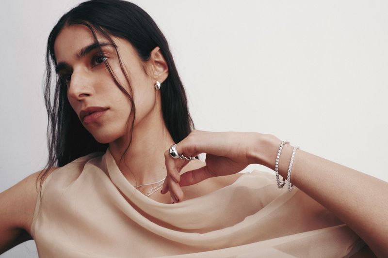 Close-up portrait of a woman wearing silver hoop earrings, rings, and tennis bracelets with a sheer beige top against a white backdrop.