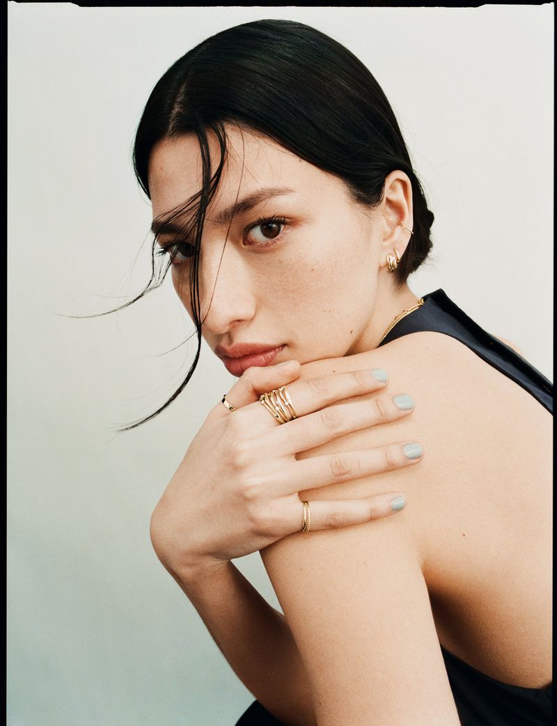 Close-up portrait of a woman wearing layered gold rings and earrings against a plain white background for Mejuri