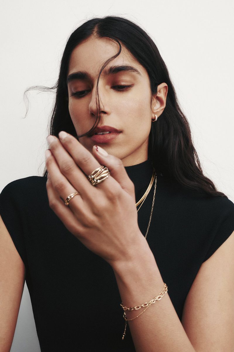 Close-up portrait of a woman wearing gold rings, necklaces, and a black top against a white seamless backdrop.