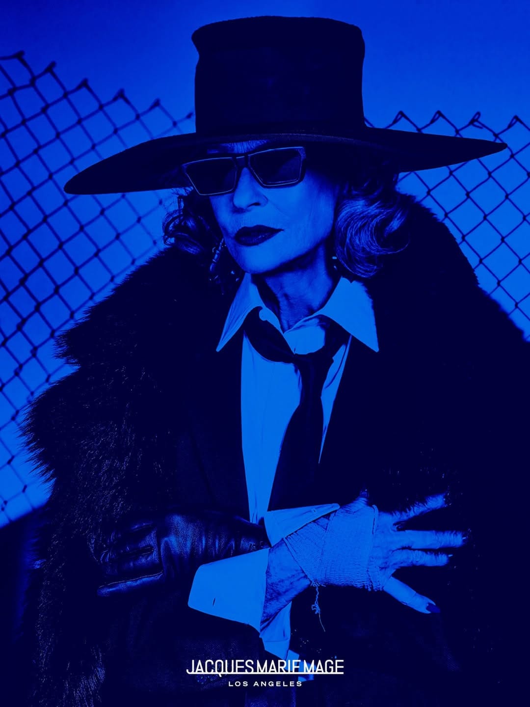 Model wearing a wide-brimmed black hat, sunglasses, white shirt, black tie, and dark coat against a chain-link fence under blue lighting.