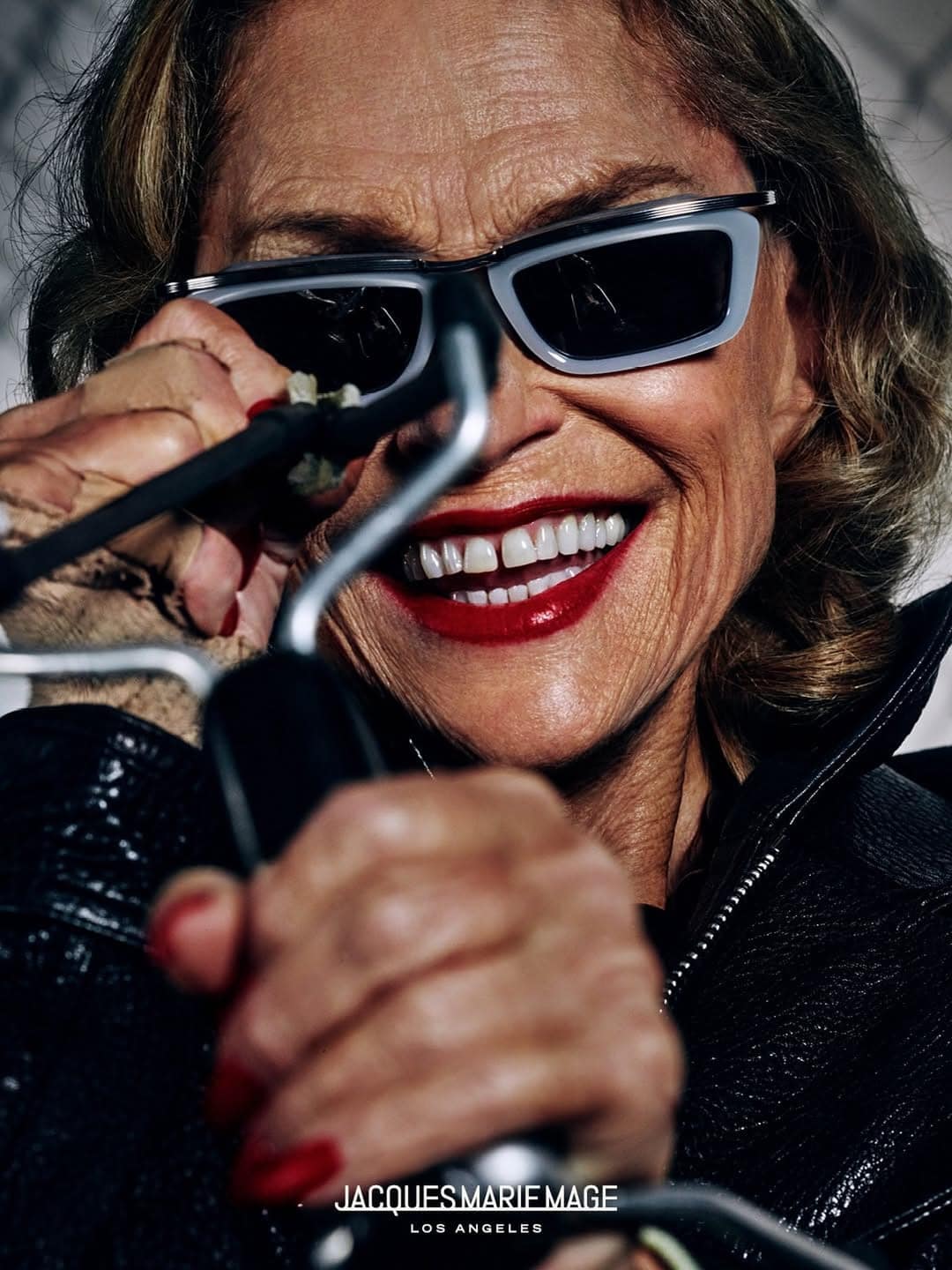 Model wearing dark sunglasses and a black leather jacket smiling broadly while holding handlebars