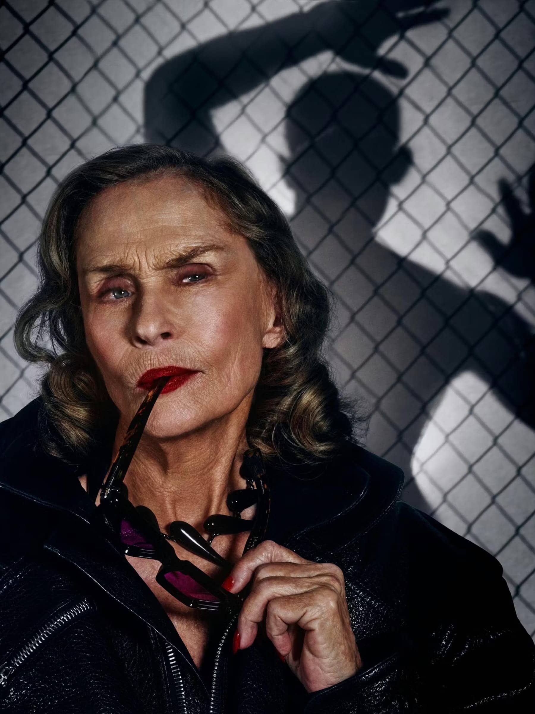 Model Lauren Hutton holding sunglasses wearing a black leather jacket against a chain-link fence shadow background