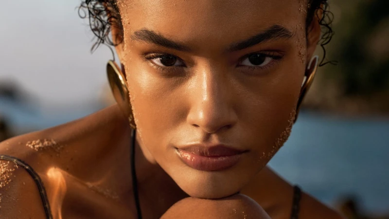 Close-up portrait of a model with glowing skin and gold earrings against a coastal background for Louis Vuitton.
