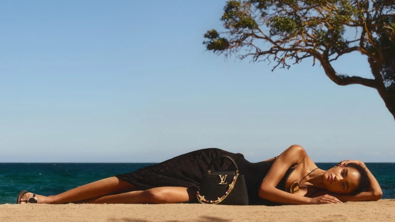 Model lying on sandy beach wearing a dark dress with a black handbag beside her under a tree