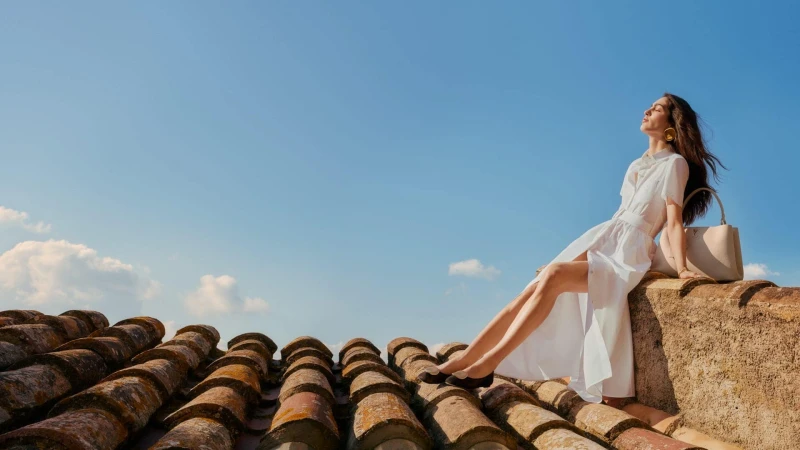 Model in white dress seated on terracotta tile rooftop against clear blue sky holding handbag