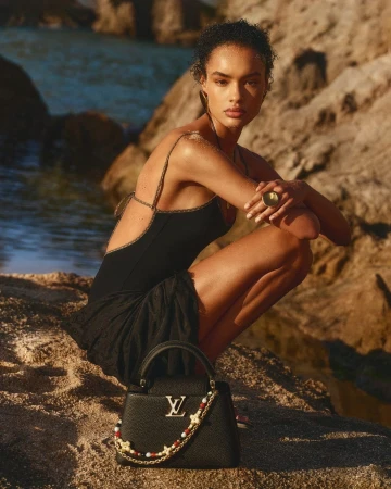 Model in black swimwear crouching on a rocky coast with a black leather handbag featuring gold Louis Vuitton hardware.