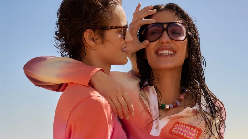 Two smiling women wearing oversized sunglasses and pink gradient polo shirts outdoors against a clear blue sky background.