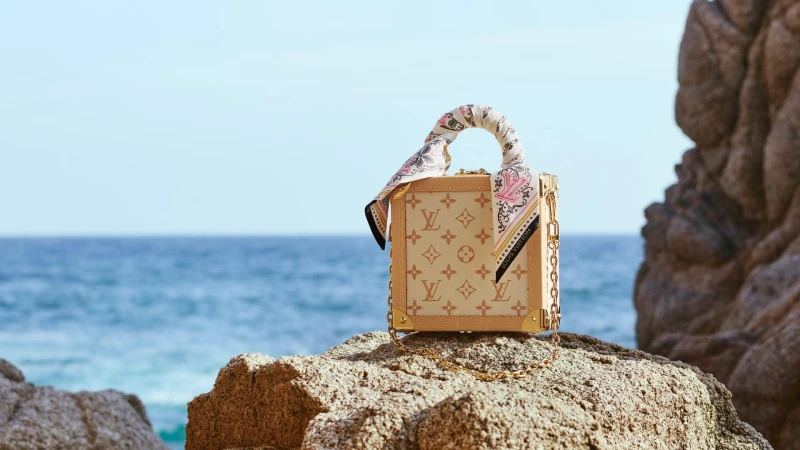 Monogram handbag with patterned silk scarf wrapped around the handle, resting on a rocky coastal shore with ocean and sky background.