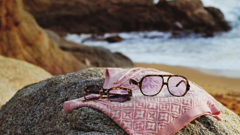 Tortoiseshell sunglasses resting on a pink patterned towel on a rock near a sandy beach and ocean waves.